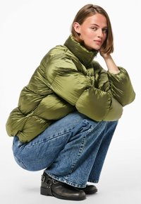 Alyvų žalia pūkinė striukė su aukštu kaklu, siuvinėtu tekstūru ir blizgiu paviršiumi, derinama su mėlynomis plačiomis kelnėmis ir juodais kulkšnies auliniais batais.