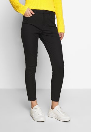 Mustat istuvat housut joustavasta kankaasta, joissa on piilotettu napillinen kiinnitys ja vinot taskut. Yhdistetty valkoisiin tennareihin.