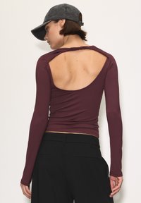 Egy nő bordó színű, hosszú ujjú felsőt visel, amely hátul kivágott, fekete nadrággal, háttal állva, sötétszürke baseballsapkával a fején.