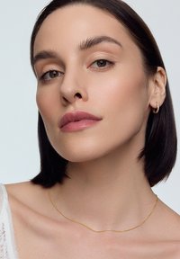 Naszyjnik w kolorze złotym z delikatnym wzorem. Modelka nosi małe, złote kolczyki w kształcie kółek. Skóra ma gładką, naturalną teksturę.