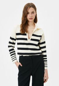 Sweter w paski z kołnierzykiem, charakteryzujący się poziomymi czarno-białymi paskami. Ma głęboki dekolt w serek oraz długie rękawy, zestawiony z ciemnymi spodniami.