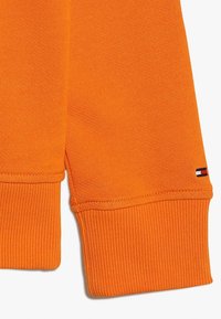 Detailní záběr na rukávy oranžové mikiny s žebrovanými manžetami a malým obdélníkovým logem v červené, bílé a námořnické modři blízko manžety.