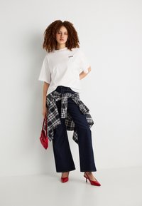 Γυναίκα με κατσαρά μαλλιά που φοράει λευκό υπερμεγέθη t-shirt, σκούρα τζιν παντελόνια με φαρδιά μπατζάκια, καρό πουκάμισο δεμένο στη μέση, κόκκινες ψηλοτάκουνες γόβες και κρατάει μια κόκκινη τσάντα.