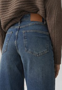 Blue denim jeans with a slightly faded texture, featuring two back pockets and a tan leather patch at the waist.