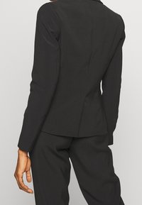 Blazer nero su misura con maniche lunghe e taglio aderente. Presenta un design minimalista, cuciture sottili e una singola fessura posteriore.