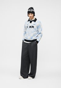 Νεαρός με μαύρο σκουφί με λευκό μοτίβο, ανοιχτό μπλε και λευκή καρό μπλούζα με κείμενο, φαρδιά σκούρα γκρι παντελόνια και λευκά παπούτσια.