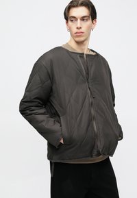 Tumepruun tikandiga bomberjakk lukuga ja küljetaskutega; ümar kaelus ja tekstuuriga materjal.