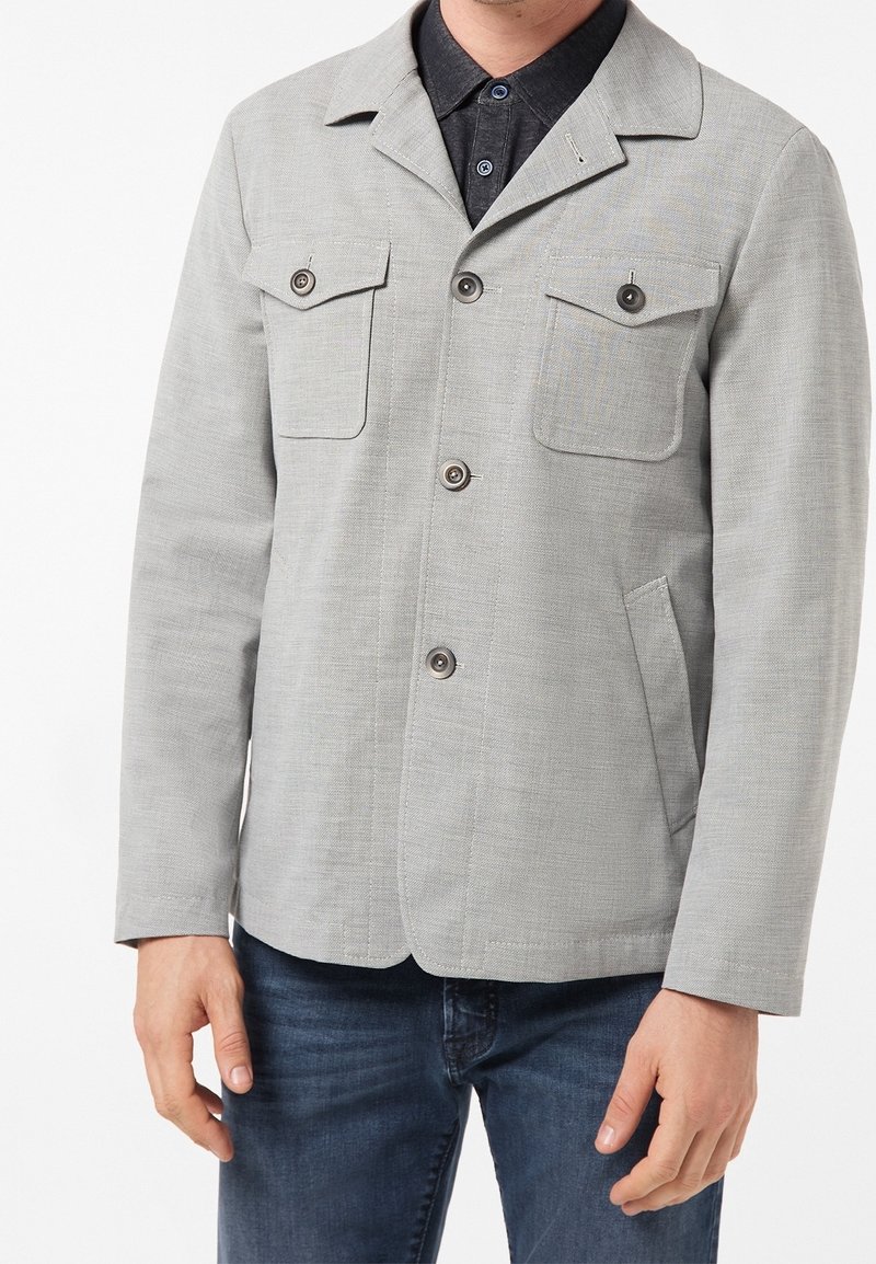 Szara, teksturowana kurtka z kołnierzem, dwoma kieszeniami na piersi i zapięciem na guziki. Noszona na ciemnej koszuli dżinsowej, w połączeniu z niebieskimi jeansami.