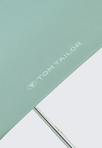 Groene paraplupunt met wit "TOM TAILOR" logo en dun zilverkleurig metalen steel tegen een lichtgrijze achtergrond.