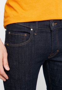Mörkblå denimjeans med slim fit, orange sömnad, framfickor och metallbeslag vid midjan och fickorna.