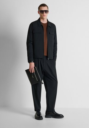 Hombre con chaqueta negra, pantalones negros, suéter marrón, gafas de sol negras y zapatos negros, sosteniendo una bolsa negra con cremallera sobre un fondo blanco.