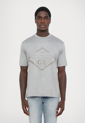 Grå bomull T-skjorte med korte ermer, med et teksturert logodesign i beige på brystet, kombinert med lyseblå jeans.