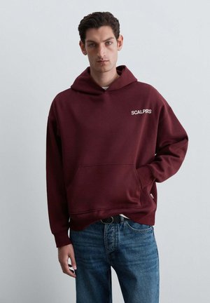 Joven con una sudadera con capucha burdeos con texto "Scalpers" y vaqueros azules, de pie frente a un fondo gris claro liso.