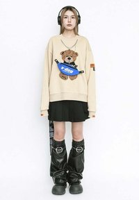 Sudadera crema con gráfico de oso de peluche, falda plisada negra, riñonera azul, pantalones baggy negros brillantes y auriculares.