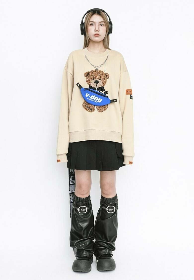 Sudadera crema con gráfico de oso de peluche, falda plisada negra, riñonera azul, pantalones baggy negros brillantes y auriculares.