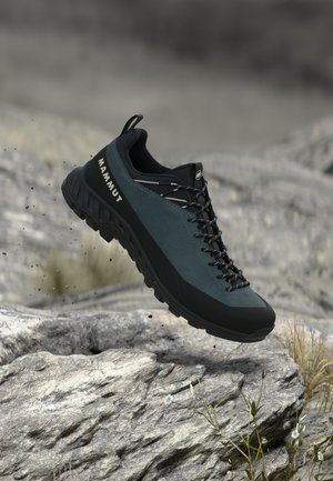 Tume roheline ja must Mammuti matkasaabas riputatud kivise maastiku kohale väikeste taimede juurde, näidates robustset tallat ja pitsi ülaosa.