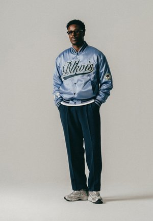 Mand iført briller, en blå satin varsity jakke med teksten "Blkvis", mørke bukser og grå sneakers, stående med hænderne i lommerne mod en ensfarvet baggrund.