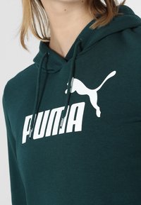 Zelená mikina s kapucí, šňůrkou, měkkou látkou a výrazným bílým logem "PUMA" s grafikou skákající kočky.