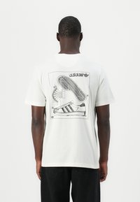 White cotton t-shirt featuring a black graphic design of a sneaker on the back, showcasing detailed schematics and the Adidas logo.