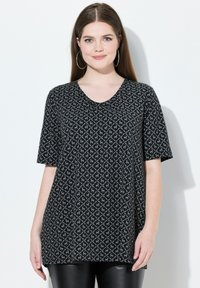 Žena s dlouhými hnědými vlasy m�á na sobě krátký rukáv s šedým a černým vzorem a velké kruhové náušnice, stojí na bílém pozadí.