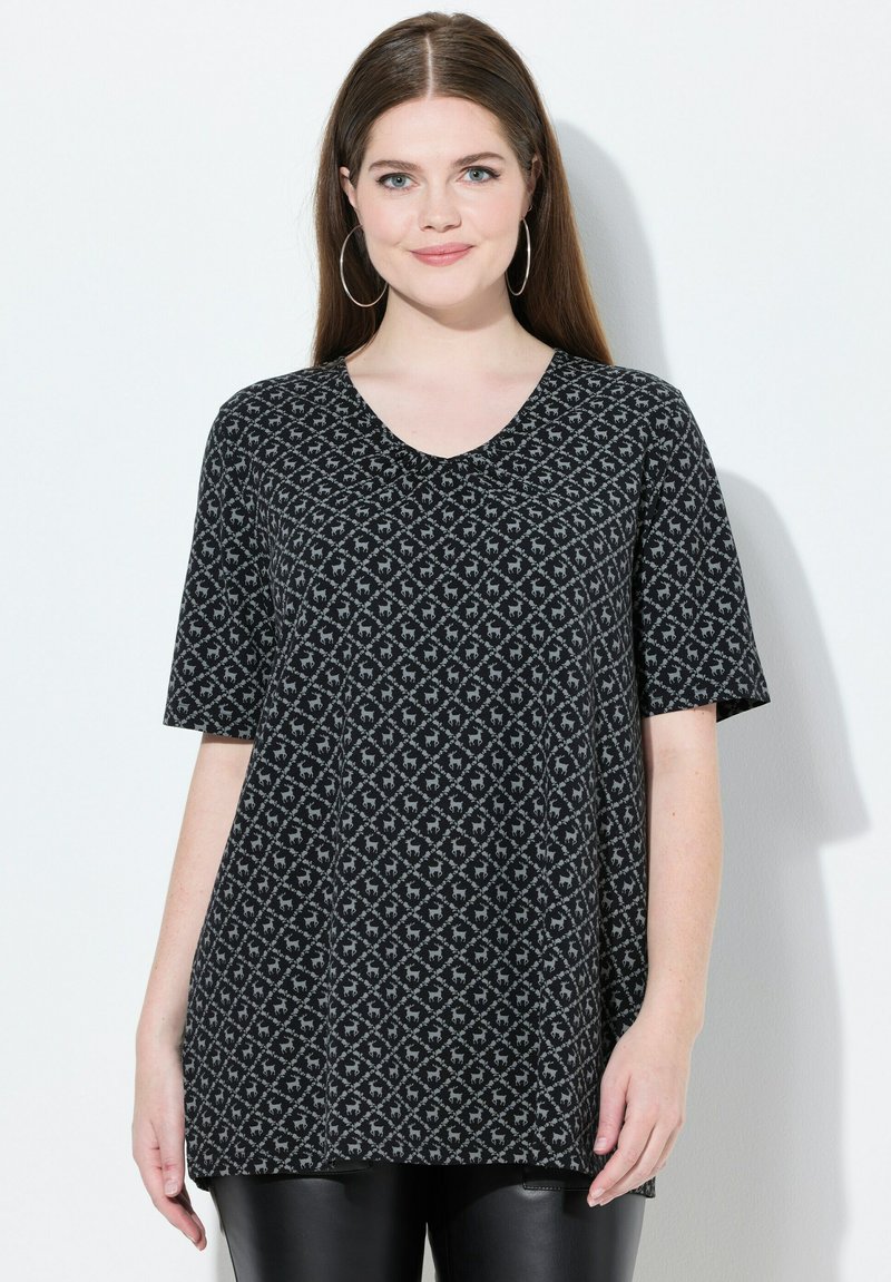 Žena s dlouhými hnědými vlasy má na sobě krátký rukáv s šedým a černým vzorem a velké kruhové náušnice, stojí na bílém pozadí.