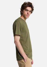 Olive green short-sleeve t-shirt with a crew neckline and a slightly textured fabric, worn with beige pants, shown in a side profile.