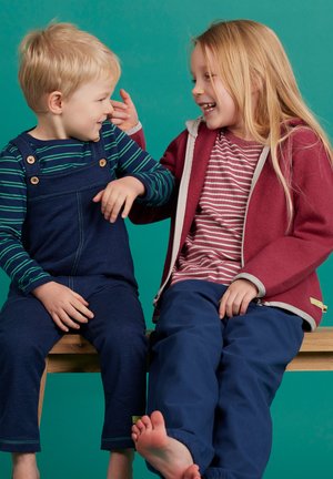 Due bambini sono seduti su una panchina di legno. Il bambino indossa una salopette blu navy con bottoni di legno e una maglietta a righe a maniche lunghe. La bambina indossa un giubbotto bordeaux con zip e dettagli grigi, insieme a una maglietta a righe rossa e bianca, abbinata a pantaloni blu.