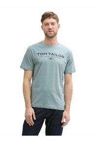 Helles blaues Baumwoll-T-Shirt mit kurzen Ärmeln, versehen mit einem dunkelblauen Logodruck auf der Brust. Getragen mit dunklen Jeans, Modell steht vor weißem Hintergrund.