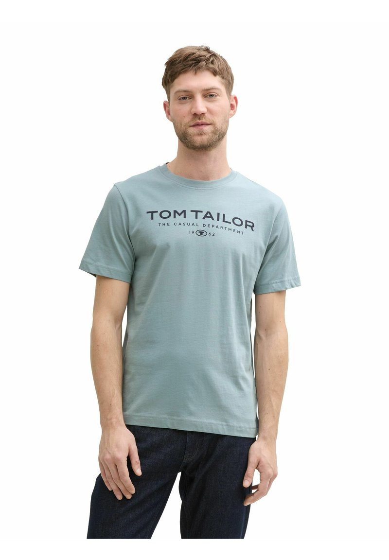 Helles blaues Baumwoll-T-Shirt mit kurzen Ärmeln, versehen mit einem dunkelblauen Logodruck auf der Brust. Getragen mit dunklen Jeans, Modell steht vor weißem Hintergrund.