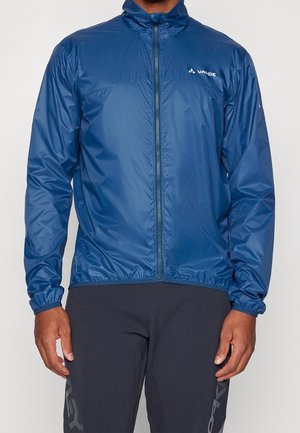 Mann trägt eine blaue Windjacke mit Reißverschluss und dem "Vaude"-Logo auf der Brust sowie schwarze Sporthose.