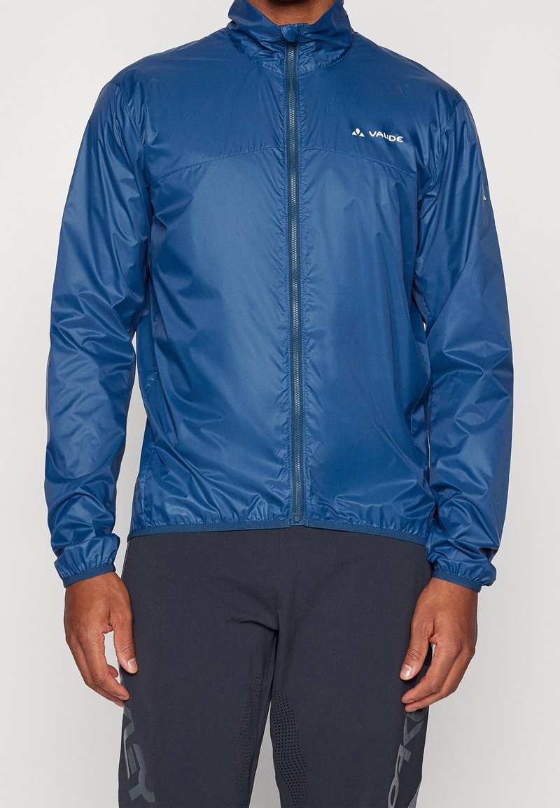 Homme portant une veste coupe-vent bleue zippée avec le logo "Vaude" sur la poitrine et un pantalon de sport noir.