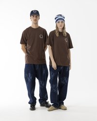 Braune Baumwoll-T-Shirts mit kleinen Brustdrucken, kombiniert mit lockeren dunklen Denim-Jeans mit blauen grafischen Mustern. Zubehör in Form von blauer Mütze und Kappe.