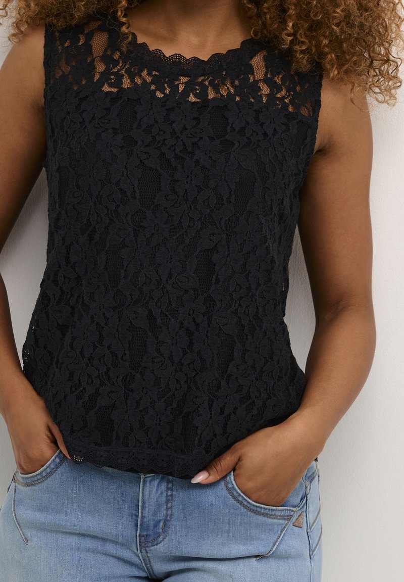 Femme portant un haut sans manches noir en dentelle florale et un jean bleu clair, les mains dans les poches, sur fond blanc.