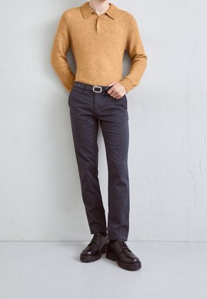 Persoon die een mosterdkleurige polo met lange mouwen draagt, donkergrijze broek met zwarte riem en zwarte veterschoenen, staand tegen een grijze muur.