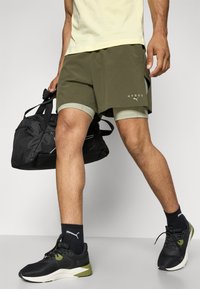 Grønne sportshorts med innvendig fôr, kombinert med en lysegul skjorte, svart sportsekk og svarte sko med grønne detaljer.