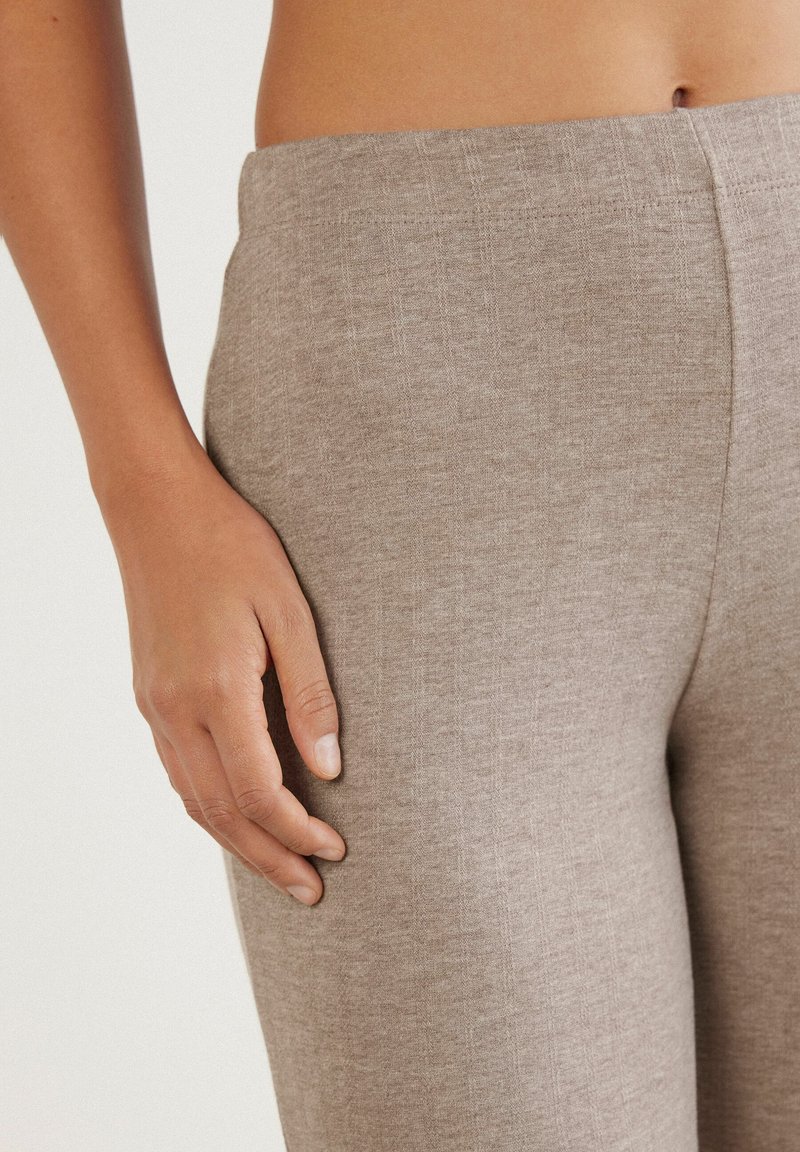Beżowe legginsy stretch z miękkiego, teksturowanego materiału, z szeroką talią i dopasowanym krojem, pokazane z ręką opartą na boku.