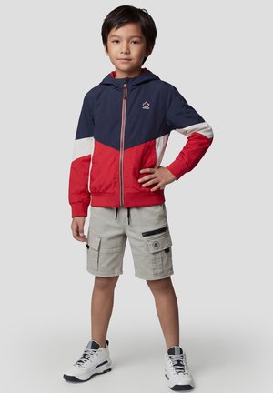 Niño de pie con la mano en la cadera, vestido con una chaqueta con cremallera roja, azul marino y blanca, pantalones cortos cargo beige y zapatillas blancas sobre un fondo gris.