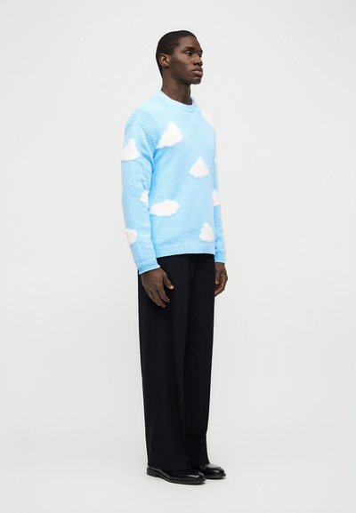 Άνδρας που στέκεται φορώντας ανοιχτό μπλε πουλόβερ με λευκά σύννεφα, μαύρο παντελόνι φαρδύ, και μαύρα παπούτσια σε απλό λευκό φόντο.