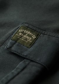 Tela gris oscuro que muestra una etiqueta textil. La etiqueta es verde con texto blanco, detallando la marca "Superdry" y la palabra "Machined".