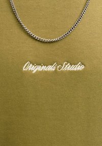 Camicia in tessuto verde oliva con finitura testurizzata, con scritta ricamata bianca "Originals Studio" e una collana di catena argentata.