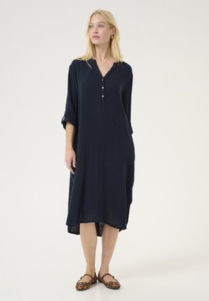 Marineblauw, knie-lange jurk gemaakt van gestructureerde stof. Heeft een V-hals, drie knopen en opgerolde mouwen, gecombineerd met leopardprint ballerina's.