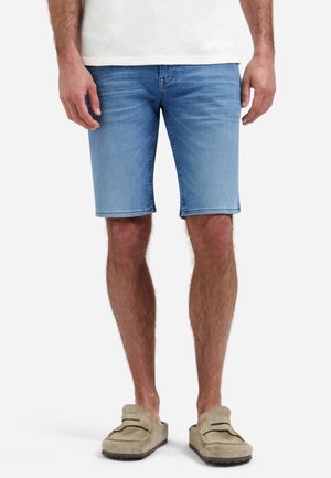 Man die lichtblauwe denim shorts, beige instapschoenen en een wit shirt draagt, staand tegen een effen achtergrond.