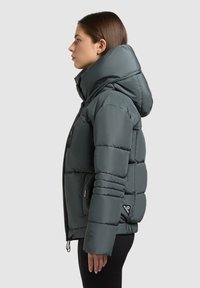 Kvinde, der står i profil iført en mørkegrøn polstret jakke med en oversized hætte og sorte bukser mod en ensfarvet grå baggrund.