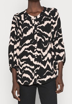 Femme portant un chemisier boutonné à motif abstrait noir et beige, avec des manches trois-quarts bouffantes, et un pantalon noir à côtes.