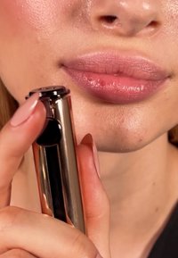 Metallisk roséguld läppstiftförpackning hålls i en hand, med en jämn, naturlig läppfärg synlig. Närbild på läppar och produkt detaljer.