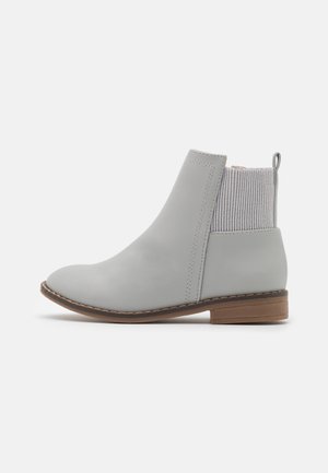 Bottine gris clair avec partie avant en cuir lisse, panneau arrière en maille côtelée, petit talon marron et languette au talon sur fond blanc.