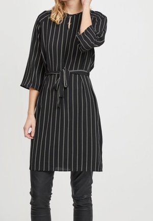 Femme portant une robe noire à rayures verticales blanches, manches trois-quarts, ceinture nouée à la taille, et pantalons en cuir noir.