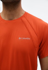 Άνδρας που φοράει αθλητικό κοντομάνικο πουκάμισο σε έντονο πορτοκαλί με διακριτική υφή πλέγματος και λευκό λογότυπο Columbia στο στήθος.