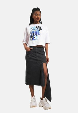 Femme portant un haut court blanc avec le texte "SOMEWHERE" et une jupe asymétrique noire avec une fente haute sur la cuisse, associée à des baskets blanches.