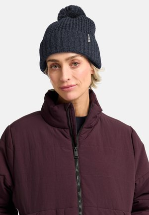 Navy knitted beanie with a pom-pom on top, paired with a maroon quilted zip jacket featuring a high collar and subtle texture.